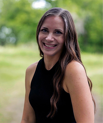 Jennifer's headshot photo outside with lots of green grass. In a black dress and her brown hair is down